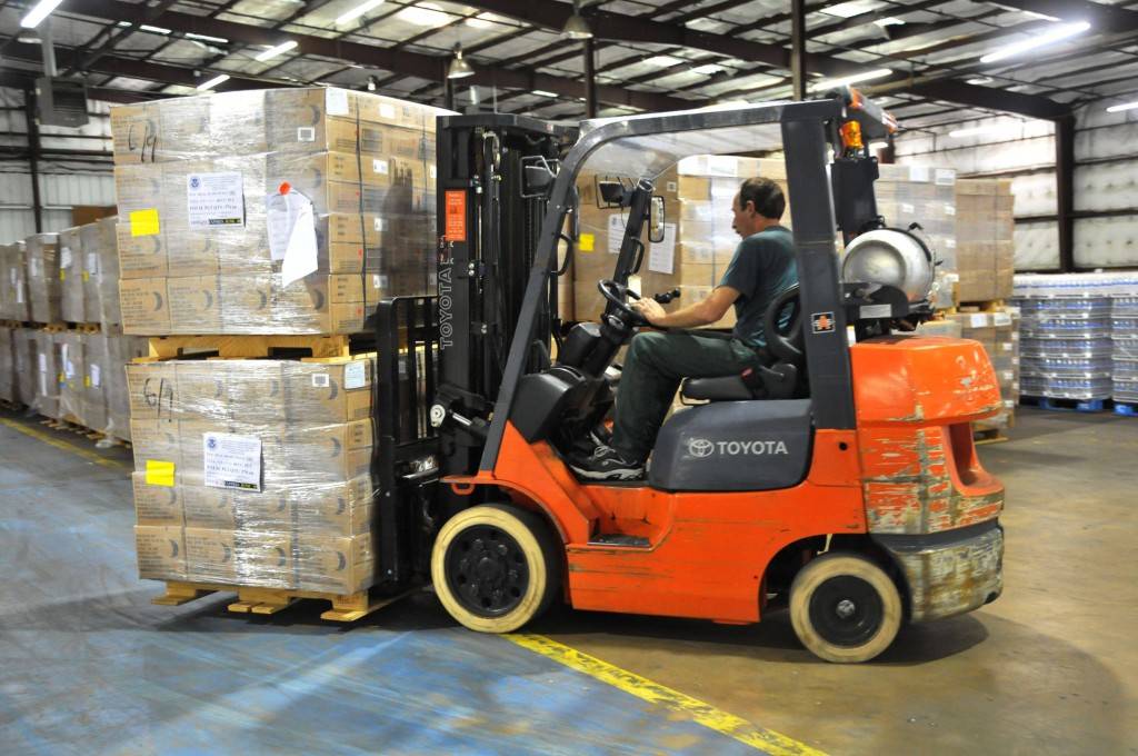 Image of a man on an orange forklift in a warehouse lifting 2 stacked pallets.
