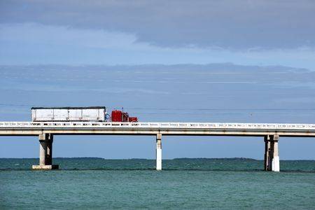 A semi truck with trailer crossing a very long bridge ove blue, blue water on a sunny day