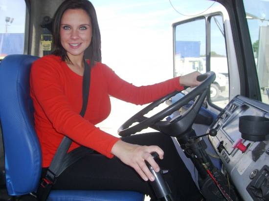 Image of a young woman driving a semi tractor trailer she is wearing a red sweater and dark pants, she has long brunette hair and a nice smile, she is facing the camera