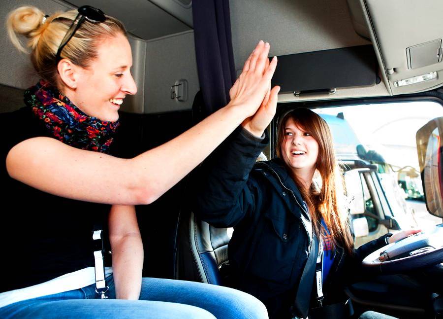 2 attractive women in the cab of a semi tractor, they are high fiving eah other, and smiling, one has long blonde hair and the other has long brunette hair.