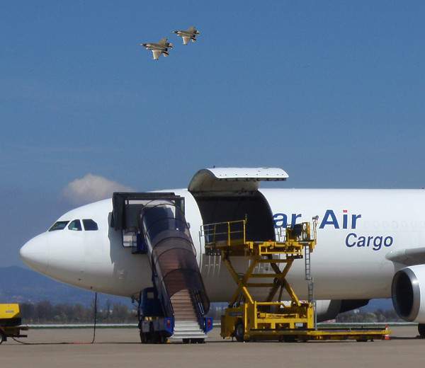 an image of a white cargo plane being loaded and there are 2 F-35 fighters doinf a high bank turn 