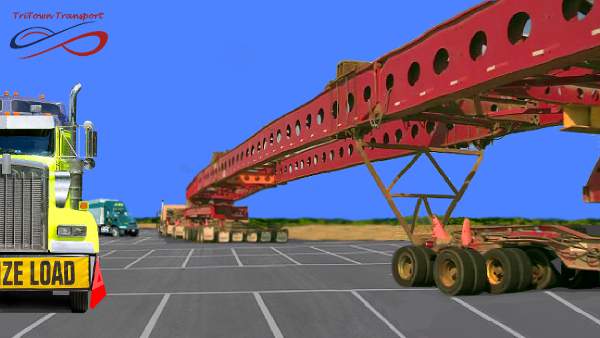 An image of an over-sized freight load, it's 2 very long metal beams or roof spans they are in a parking lot and the tractor is in the distance and the span is close to the camera 