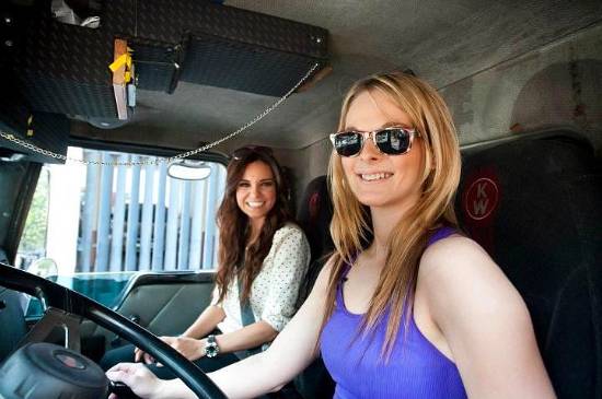 an image of 2 attractive semi truck drivers in the cab of a truck one has long blonde hair and one woman has brunette hair the blonde is wearing sunglasses they are both wearing tank tops and smiling at the camera