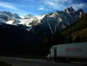 a beautiful image of a specialized freight truck hauling a load thru a mountian pass, its a summerday with pine trees below and towing mountians in the background