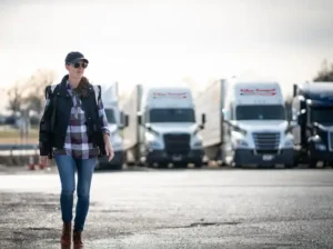 AN image of a woman trucker walking aross the street at a truck stop cafe, she has 3 tritown transport truck in the background, she look happy with a big smile for the camera