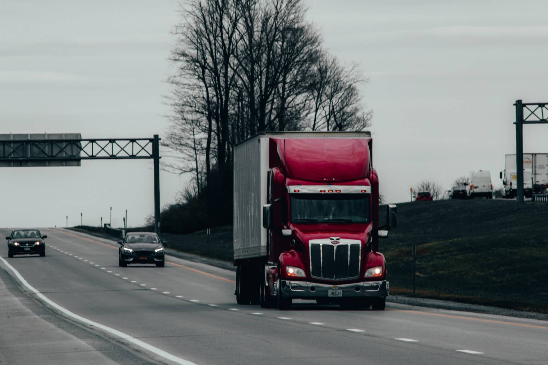 A red semi-tractor-trailer truck on the highway with a few other passenger cars. The weather is overcast but the road is dry and clear.