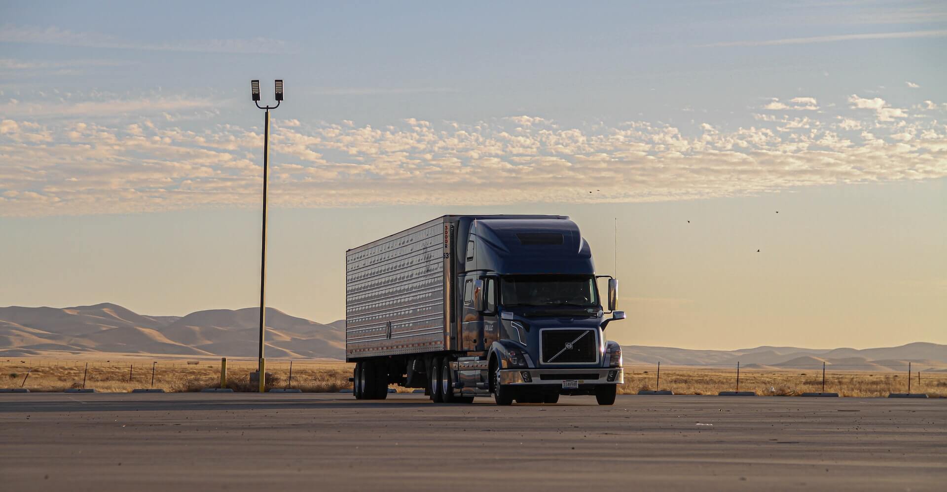 A freight truck with trailer parked in an empty parking lot just before dusk with low rolling brown hills in the distance.