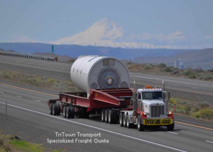 an image of a man of color driving a specialized freight load on a flatbed trailer. It's a very large piece of power generation equipment. It looks like a cool autumn day somewhere east of the rocky mountains which are in the distant background.
